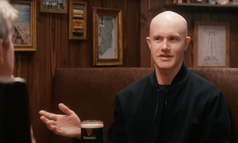 Bald man in black jacket speaks, Guinness pint nearby; warm pub setting.