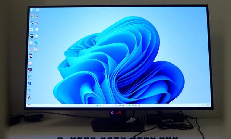 Windows 11 desktop displayed on a monitor, showing a blue abstract wallpaper and various app icons.