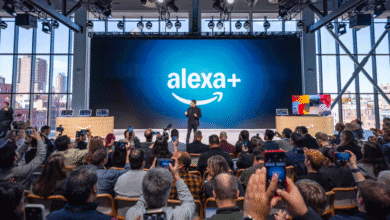 A speaker presents at an Amazon Alexa+ event, a large audience holds up phones to record.