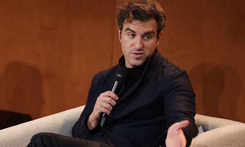 Brian Chesky, Airbnb CEO, speaks at a microphone during an event. He's seated and gesturing.