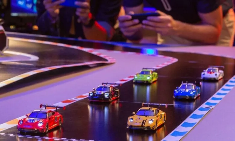 Miniature Porsche race cars compete on a tabletop track, watched by onlookers.