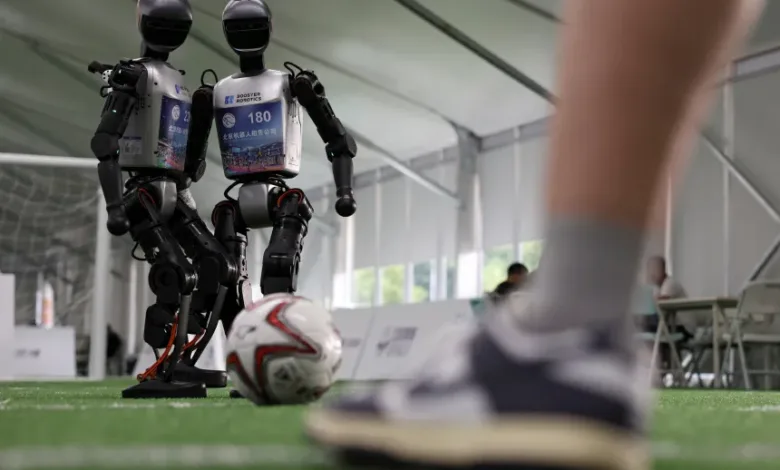 Two humanoid robots, numbered 12 and 180, play soccer on an indoor turf field. A person's legs and feet are visible in the foreground.