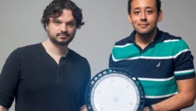 Two men hold a large, round LED light. One wears a black shirt, the other a teal and navy striped polo.