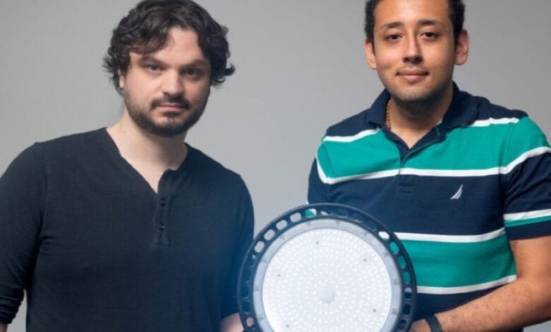 Two men hold a large, round LED light. One wears a black shirt, the other a teal and navy striped polo.