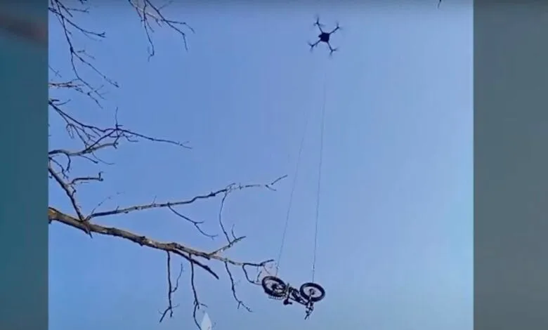 Drone lifts a bicycle suspended from a thin rope against a clear blue sky and bare tree branches.