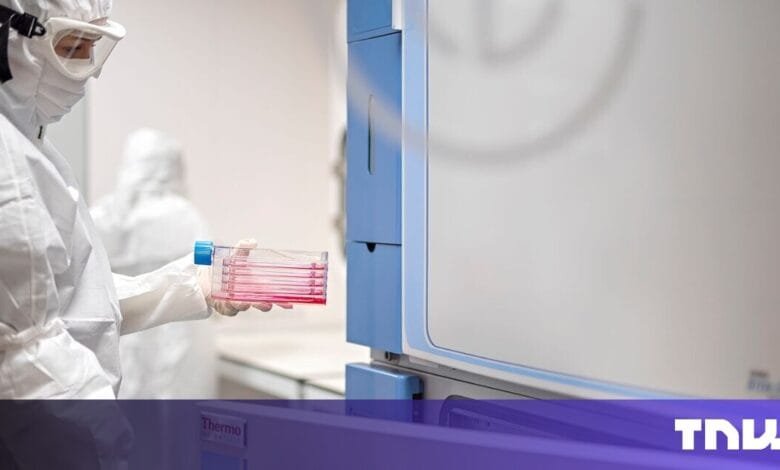 Scientist in PPE inserts sample plates into lab equipment. Red liquid is visible.