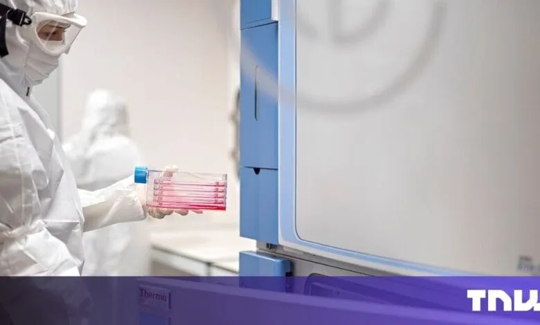 Scientist in PPE inserts sample plates into lab equipment. Red liquid is visible.