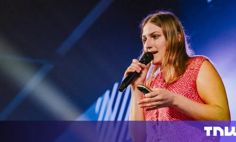 Woman speaks into microphone at a conference, holding a small device. Stage lighting creates a dramatic effect.