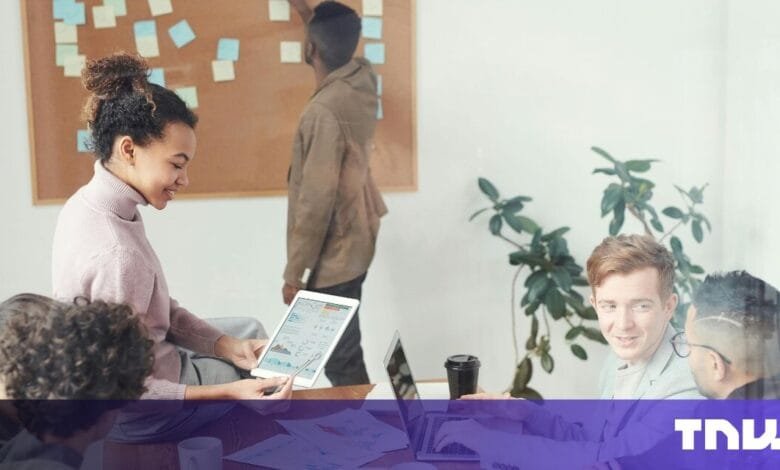 Diverse team collaborates in modern office, using laptops and tablets. A whiteboard with sticky notes is in the background.