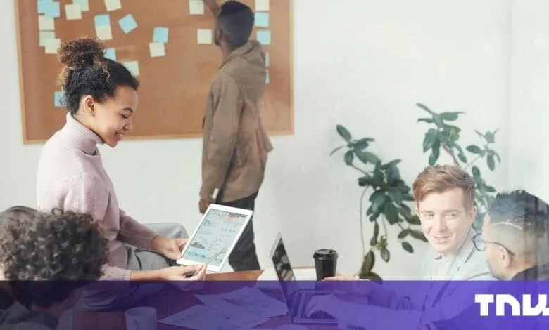 Diverse team collaborates in modern office, using laptops and tablets. A whiteboard with sticky notes is in the background.