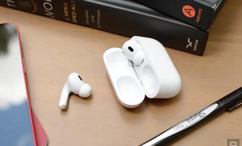 White Apple AirPods and charging case rest on a wooden desk near books and a pen.