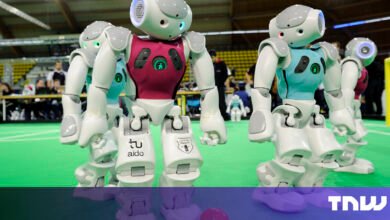 Humanoid robots in a soccer match, wearing teal and maroon jerseys. They stand on a green field.