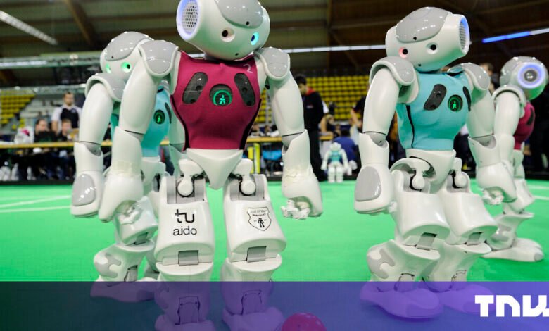Humanoid robots in a soccer match, wearing teal and maroon jerseys. They stand on a green field.