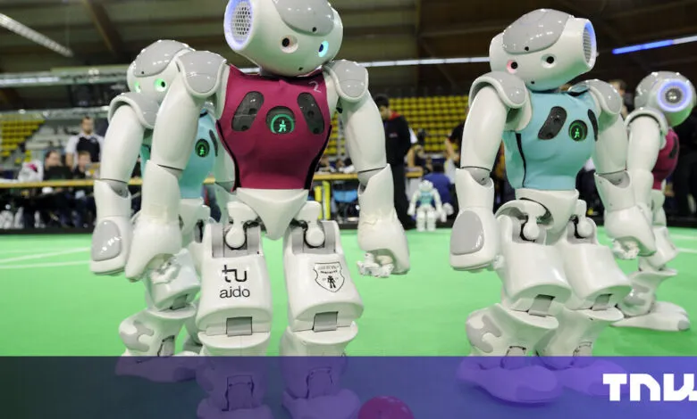 Humanoid robots in a soccer match, wearing teal and maroon jerseys. They stand on a green field.