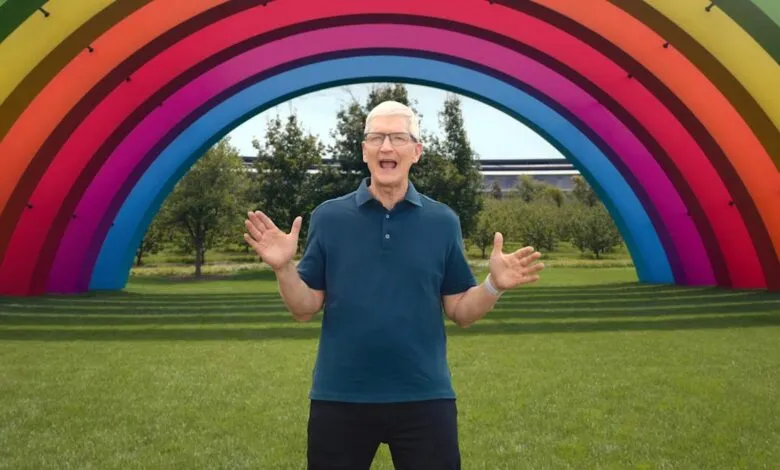 Tim Cook, Apple CEO, stands joyfully before a large rainbow arch in a park.