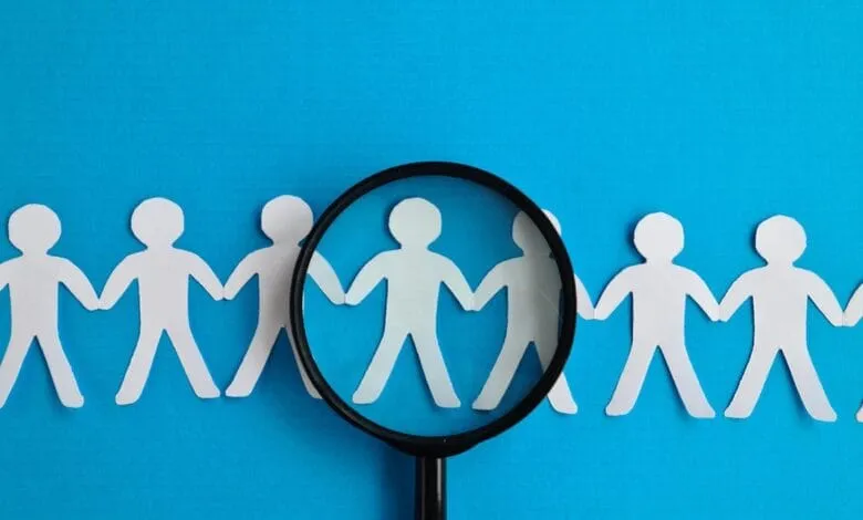Magnifying glass focuses on a single paper person in a line of linked figures, symbolizing talent search or individual selection.