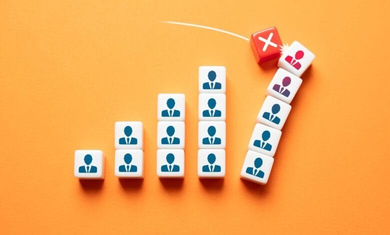 White blocks with person icons form a bar graph, a red block with an 'X' knocks one off, symbolizing employee reduction.