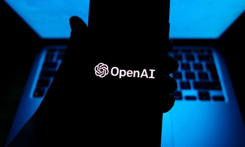 Silhouetted hand holds phone displaying the OpenAI logo against a blurred blue-lit laptop.