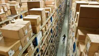 High-angle view of a warehouse aisle with towering shelves packed with cardboard boxes; a person inspects inventory.