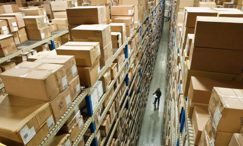 High-angle view of a warehouse aisle with towering shelves packed with cardboard boxes; a person inspects inventory.