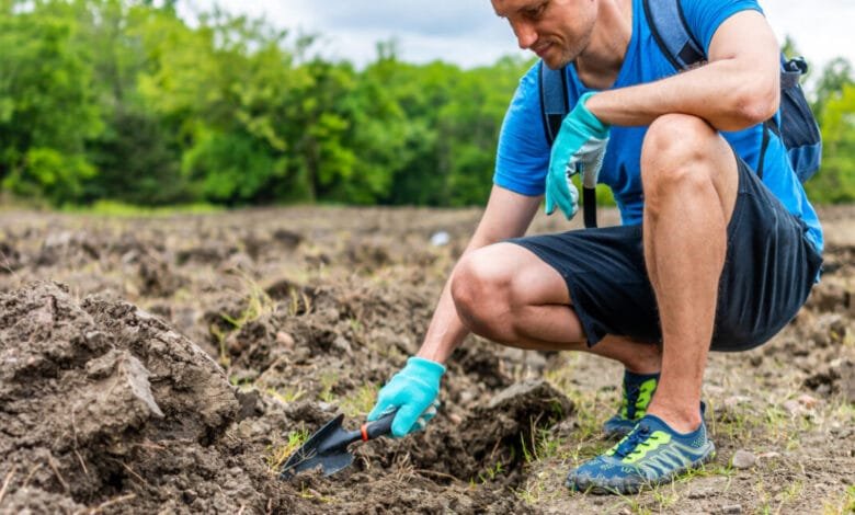 Man gardening, kneeling in a field, using a small trowel. He wears blue shirt, dark shorts, and teal gloves.