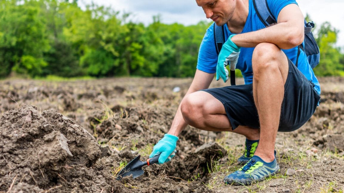 Man gardening, kneeling in a field, using a small trowel. He wears blue shirt, dark shorts, and teal gloves.