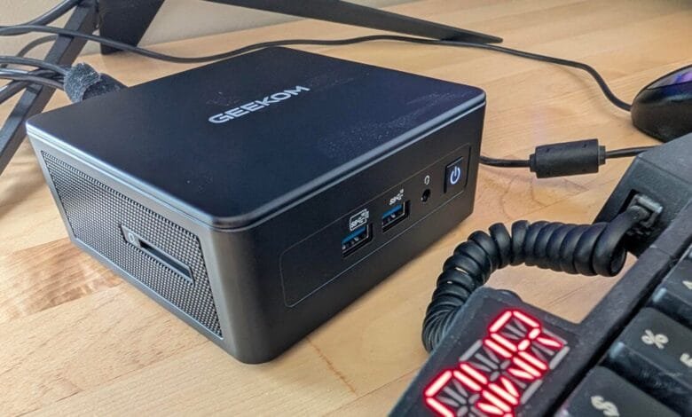 A black Geeekom mini PC sits on a light wood desk next to a keyboard and mouse. Various cables are visible.