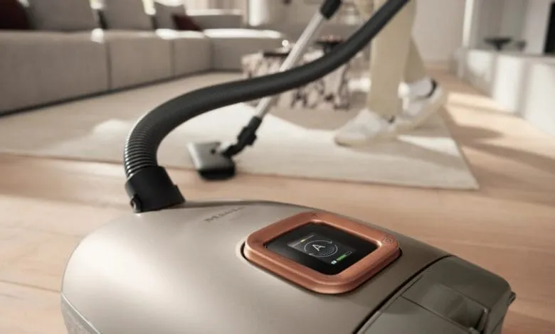 Close-up of a Miele vacuum cleaner's control panel, with the hose extending into a blurred background of a living room.