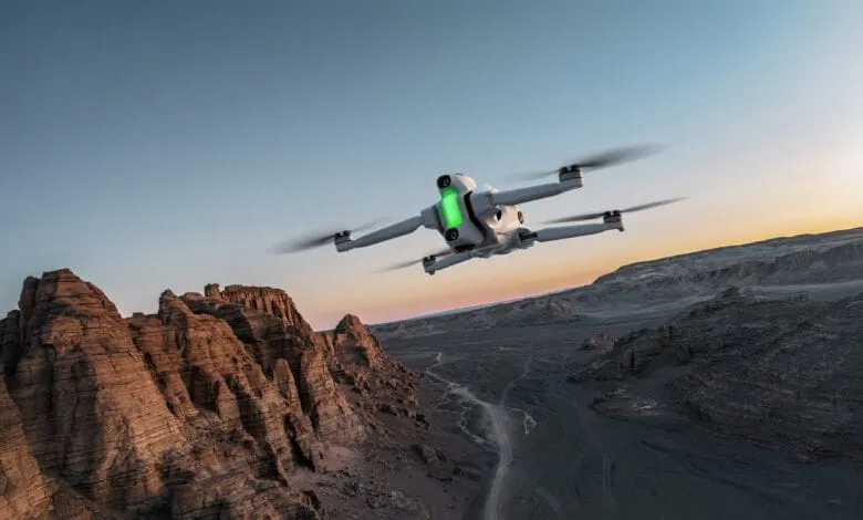A white drone with a green light flies over a desert landscape at sunset.