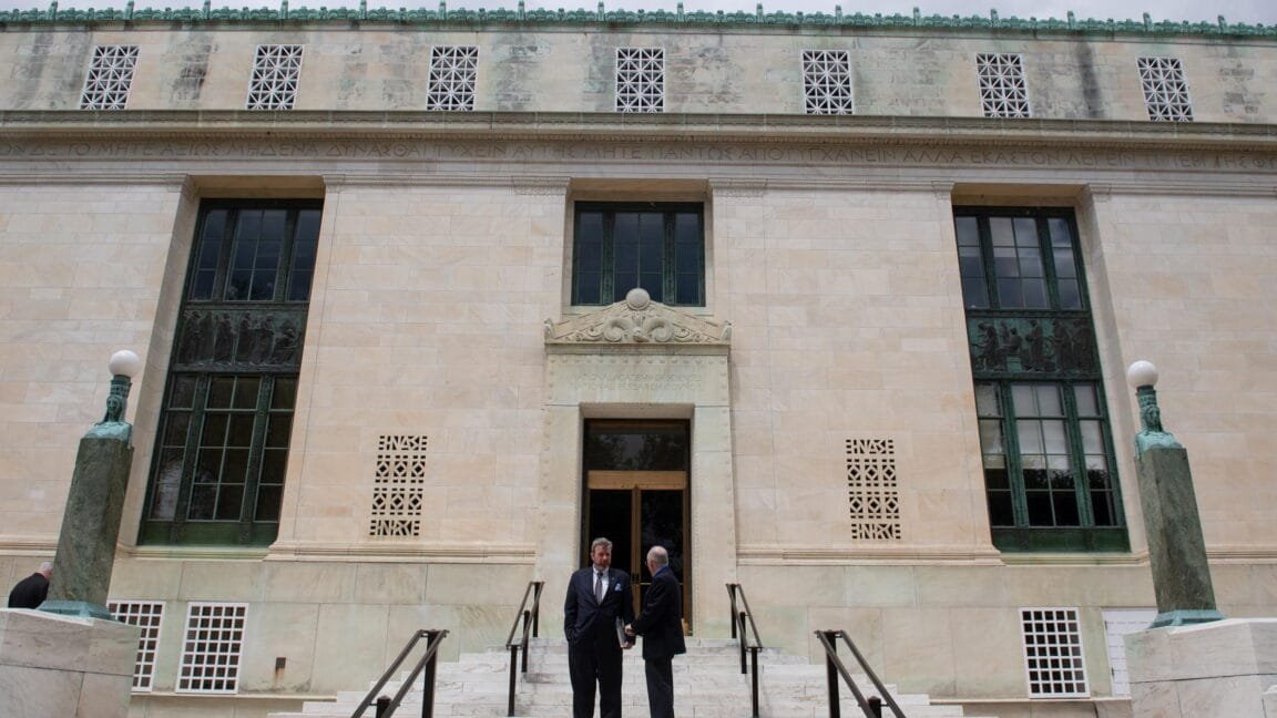 Two men in suits converse on the steps of a grand, light-beige stone building with intricate detailing and Greek inscriptions.