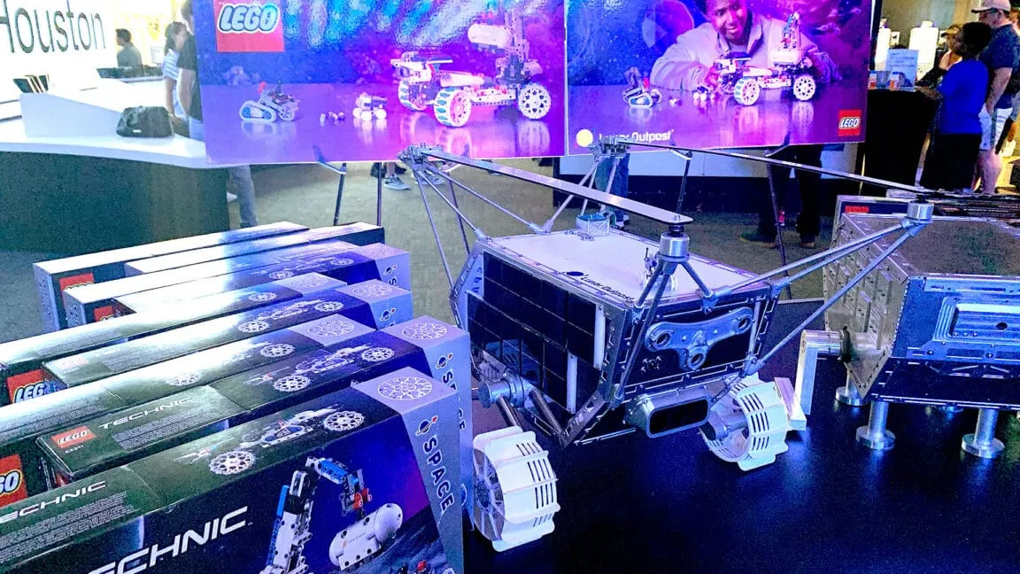 A detailed model of a lunar rover sits on display, surrounded by boxes of LEGO Technic Space sets. Purple lighting enhances the scene.