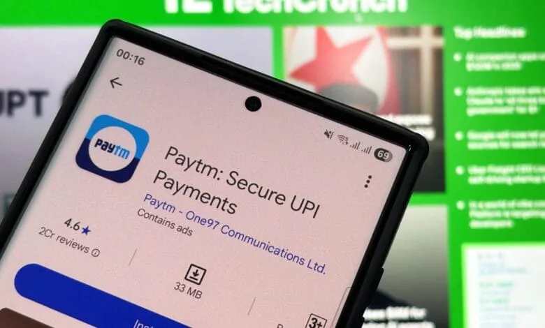 Close-up of a smartphone displaying the Paytm app on the Google Play Store, with a blurred TechCrunch website in the background.