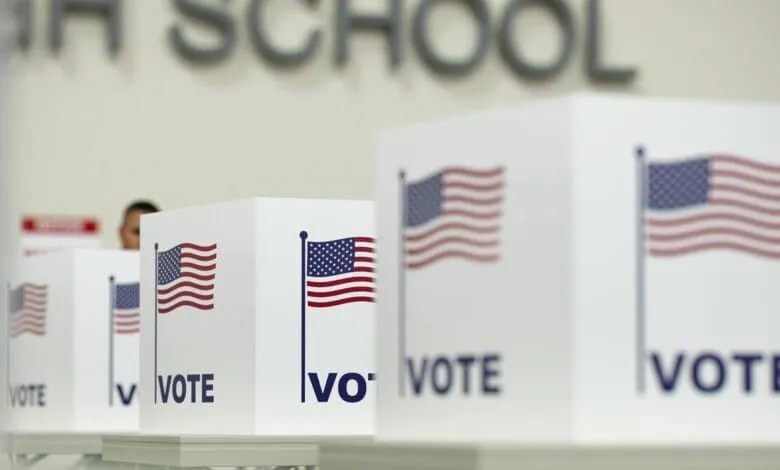 Close-up of several white ballot boxes with American flags and 'VOTE' printed on them, set against a blurred background.