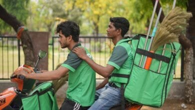 Two workers in green uniforms ride a scooter, carrying brooms in large green backpacks.