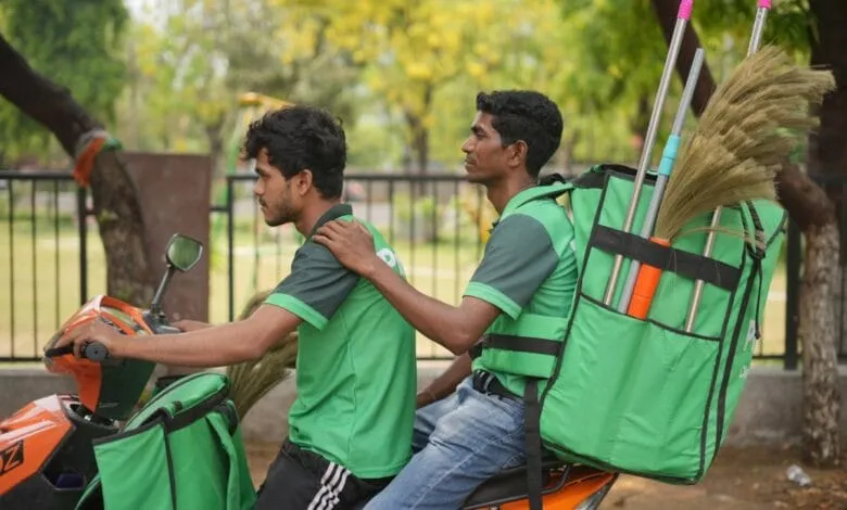 Two workers in green uniforms ride a scooter, carrying brooms in large green backpacks.