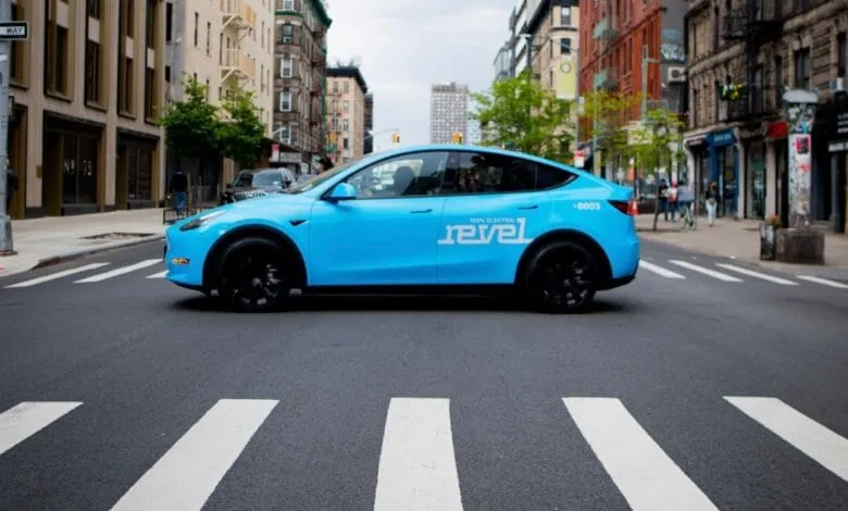 A bright blue Tesla Model Y, branded for Revel rideshare, drives across a city street.
