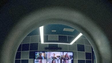 A video conference displayed on a screen in a modern room, viewed through a curved archway. The room has teal walls and bright lighting.