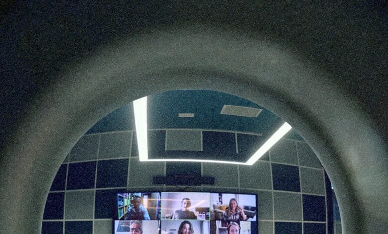 A video conference displayed on a screen in a modern room, viewed through a curved archway. The room has teal walls and bright lighting.