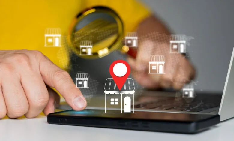 Person using phone and laptop to locate businesses on a map, using a magnifying glass to zoom in on a location.