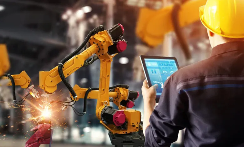 Factory worker monitors robotic arms welding a car part using a tablet.