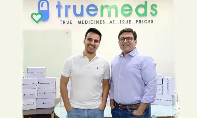 Two men stand smiling in front of a Truemeds sign, boxes of Truemeds products are stacked nearby.