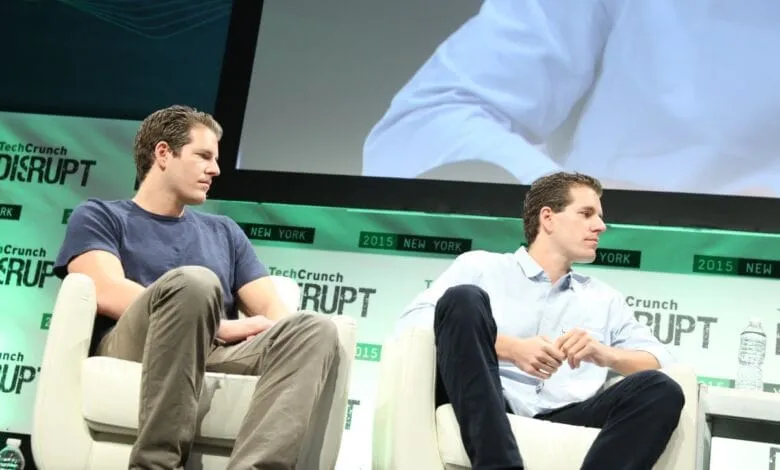 The Winklevoss twins, Tyler and Cameron, sit in chairs on a stage at a TechCrunch Disrupt conference.