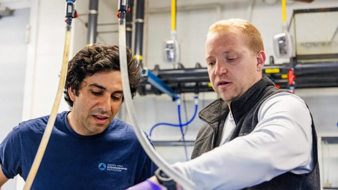 Two scientists collaborate on equipment in a lab, examining tubing and apparatus.