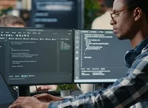 Focused programmer working on code on dual monitors in a busy office.