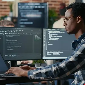 Focused programmer working on code on dual monitors in a busy office.
