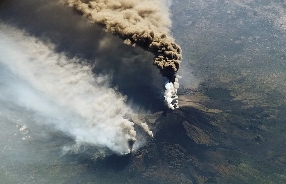 Aerial view of a volcano erupting, plumes of ash and smoke billowing into the sky.
