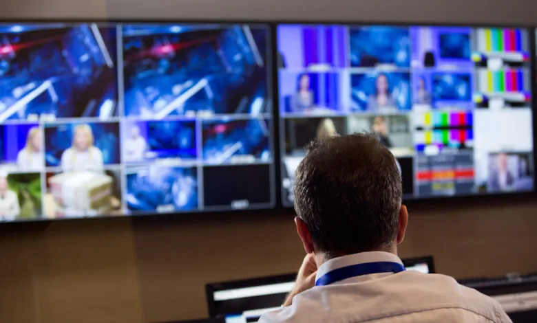 A person monitors multiple video feeds on large screens in a control room.