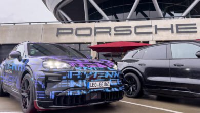 A camouflaged Porsche Cayenne prototype with a colorful wrap and a black Cayenne parked outside the Porsche building.