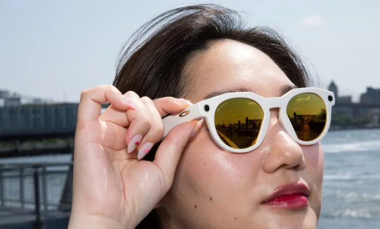 Woman adjusts white Oakley smart glasses with gold-tinted lenses, reflecting a cityscape.