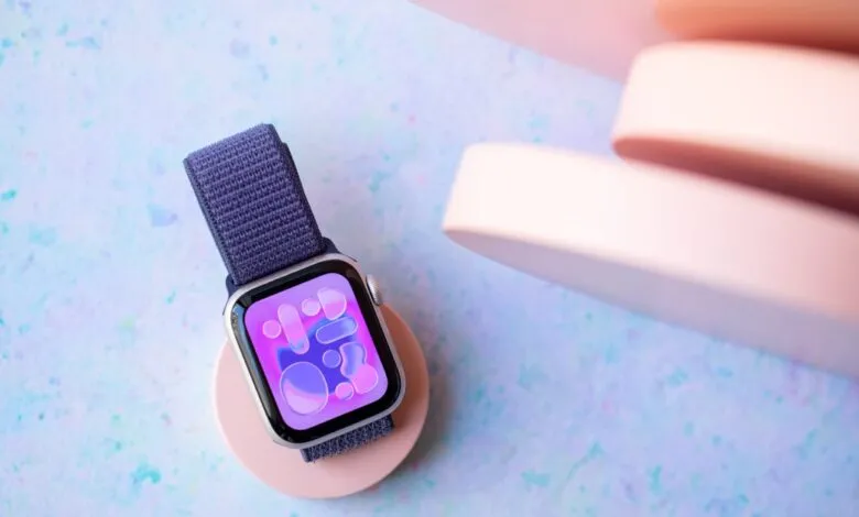 Apple Watch SE on a pink charger, displaying an abstract watch face. Soft blue background.